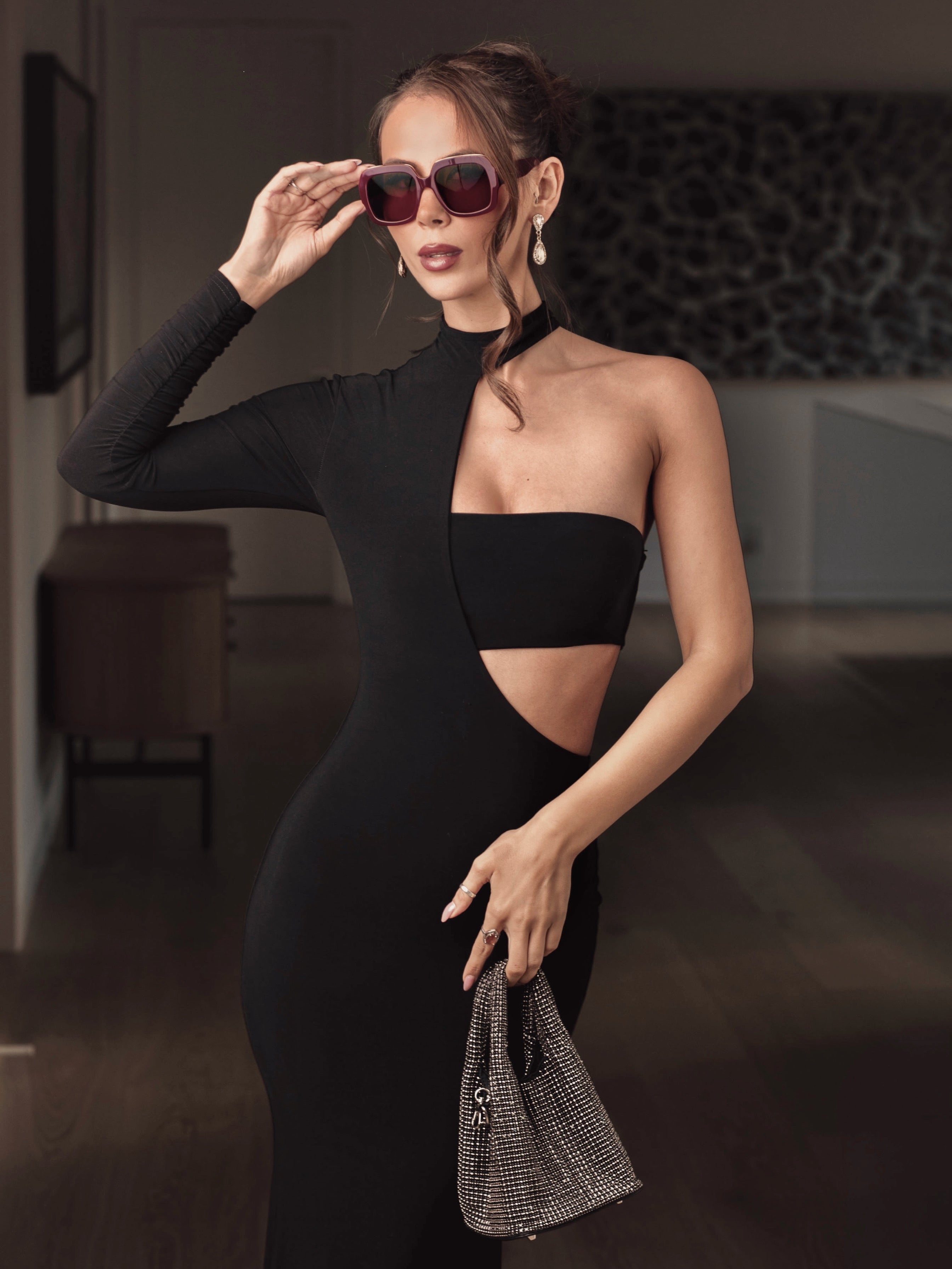 Woman in a black dress holding sunglasses and a handbag indoors