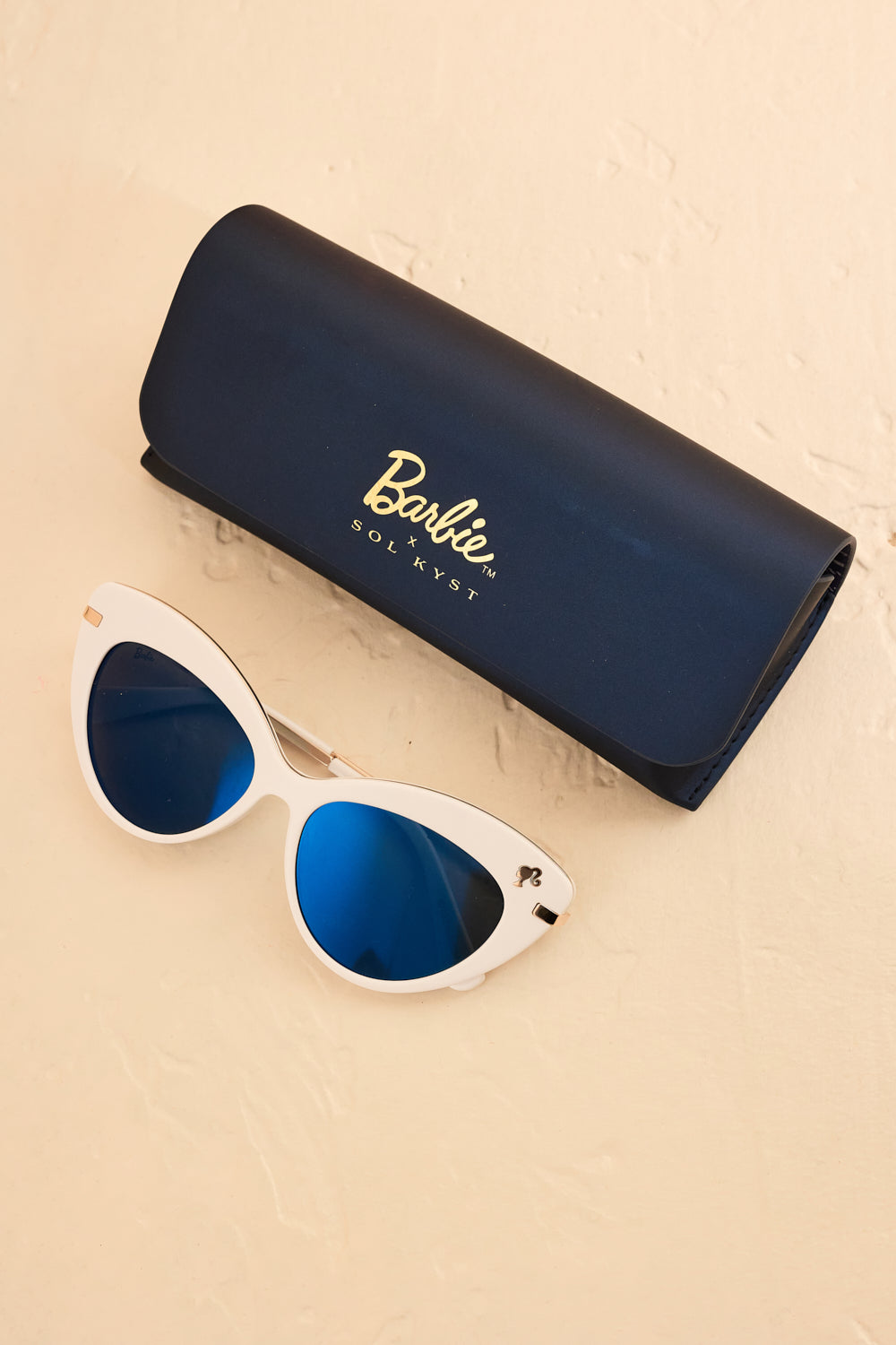 White Barbie cat eye sunglasses with blue lenses next to matching "Barbie x SOL KYST" case.