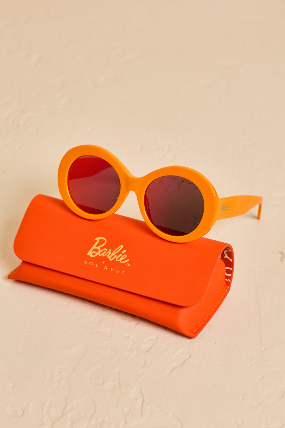 Barbie Sol Kyst round orange sunglasses with red-tinted lenses, displayed on top of an orange case on a textured beige surface.