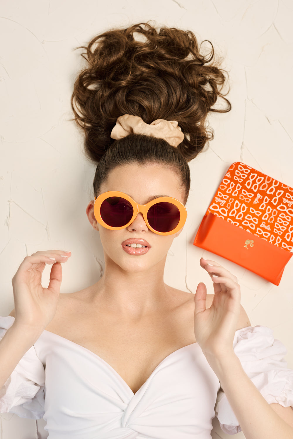 Model wearing white dress with Barbie Sol Kyst round sunglasses with orange frames next to matching case.