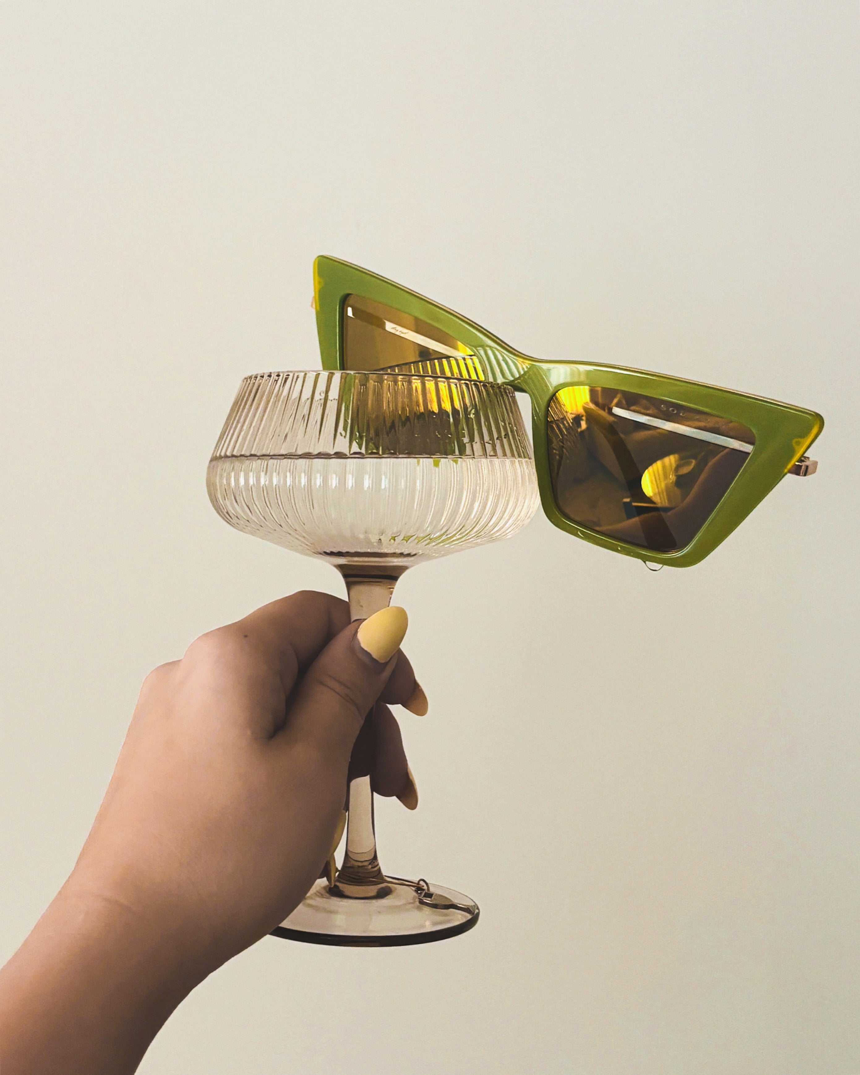 Women holding coupe glass with Sol Kyst green trapezoid sunglasses resting on the rim.