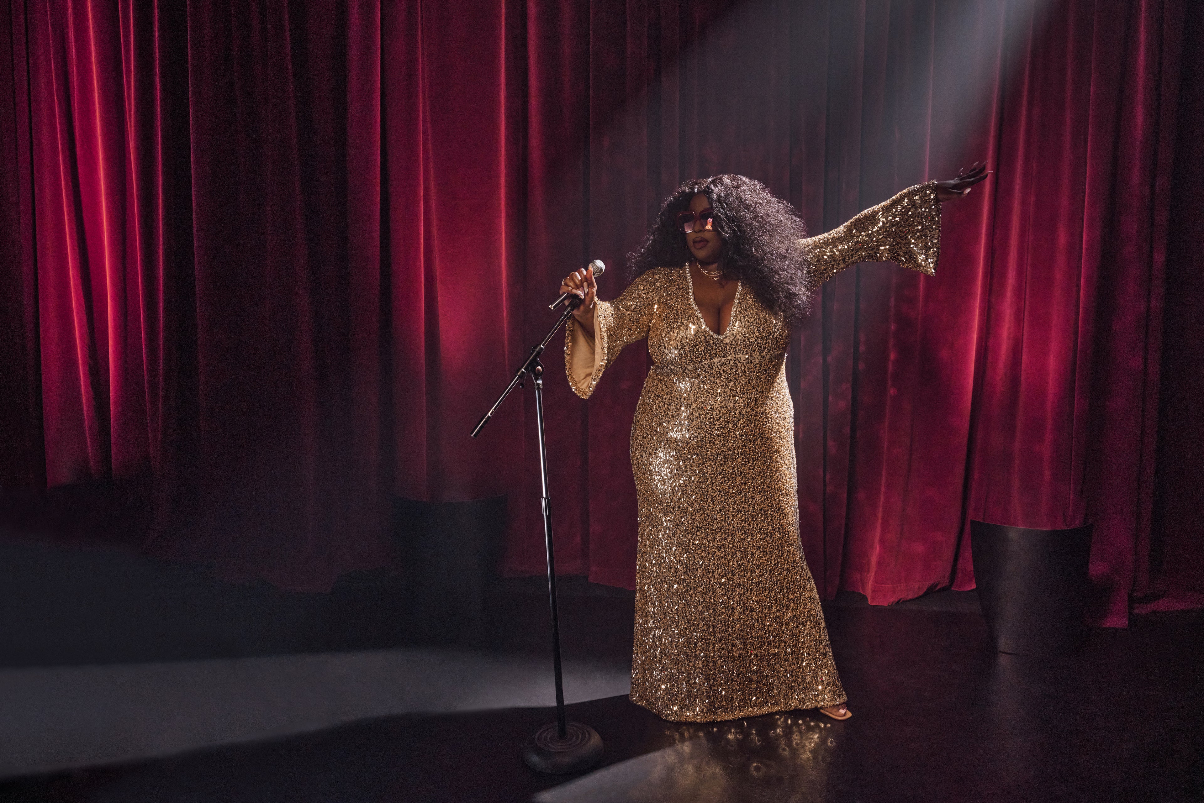 Singer standing on stage wearing shiny gold dress, wearing Barbie Sol Kyst sunglasses.