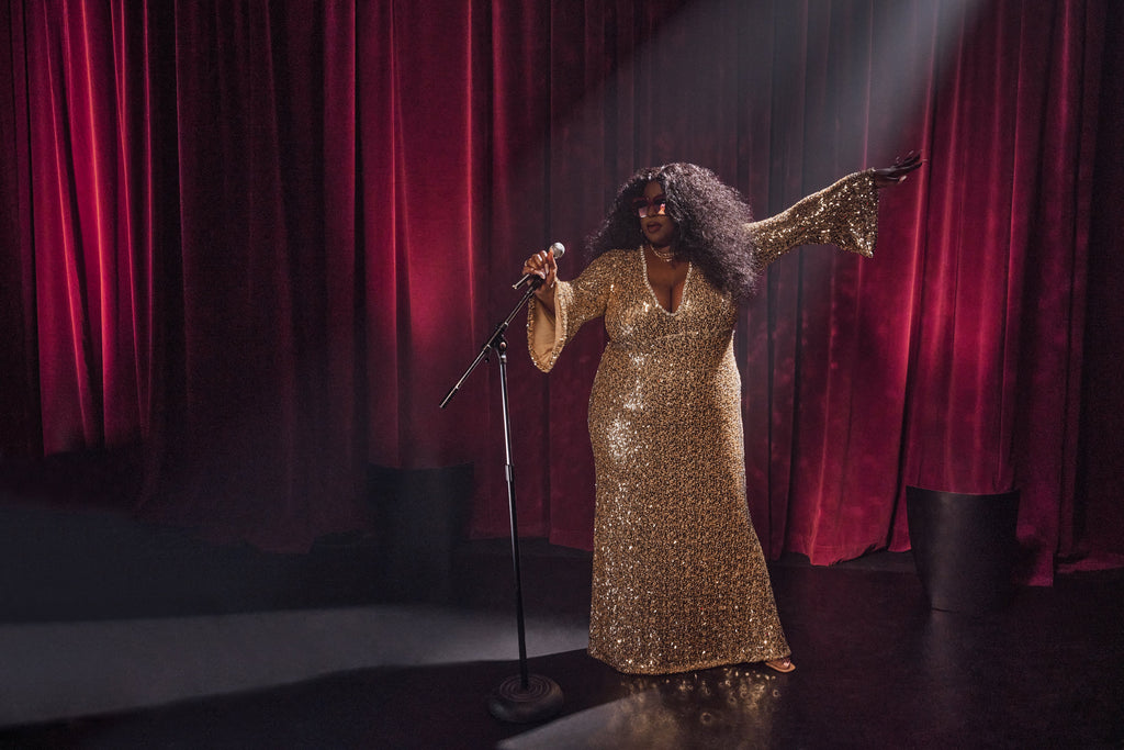 Singer standing on stage wearing shiny gold dress, wearing Barbie Sol Kyst sunglasses.