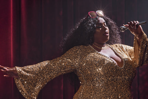 Close-up of a performer in a gold sequined outfit, with Barbie Sol Kyst square sunglasses on her head, singing on stage.