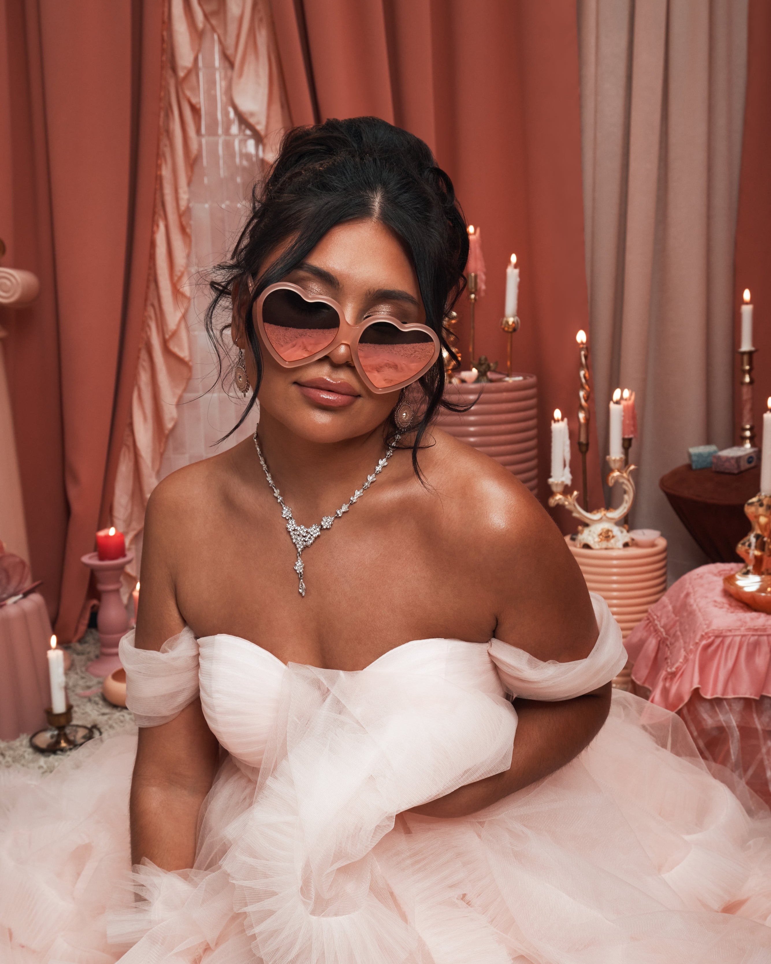 Woman in a soft pink tulle gown wearing Barbie x Sol Kyst heart-shaped sunglasses, surrounded by peach-toned decor and candles.