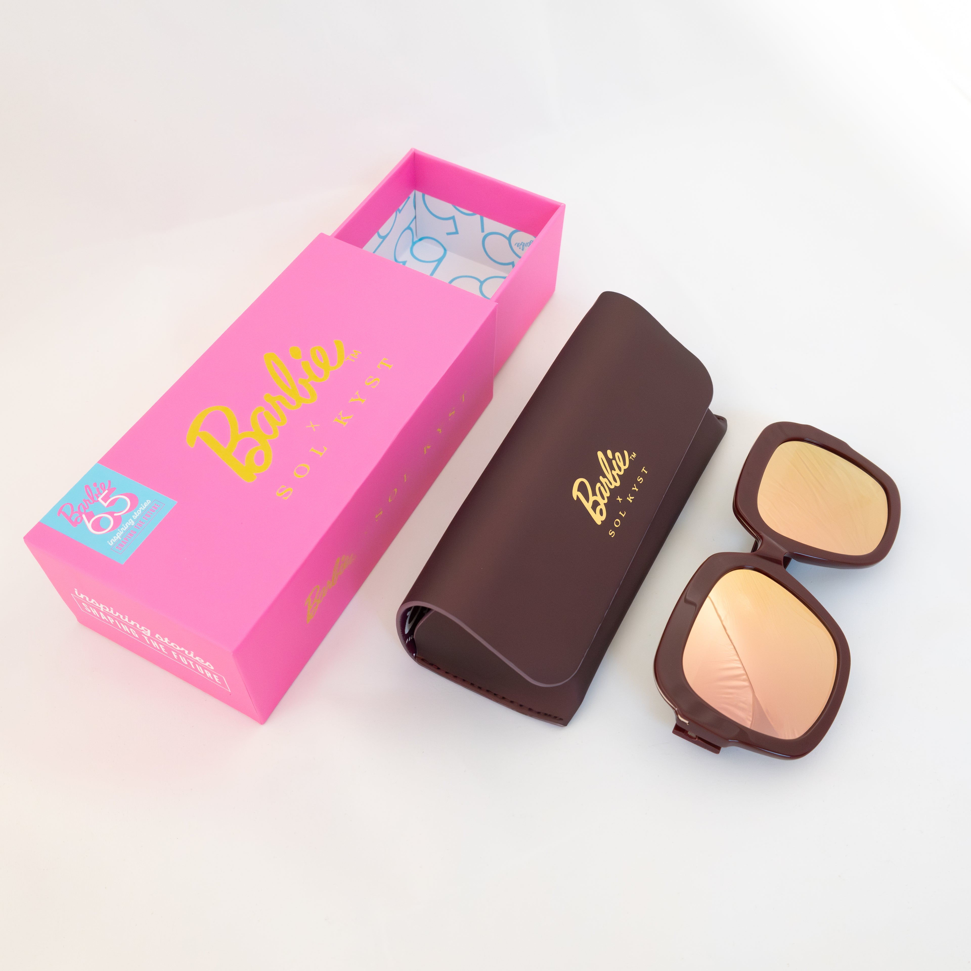 Barbie Sol Kyst sunglasses with burgundy frames and gold reflective lenses, displayed with matching case and pink box.