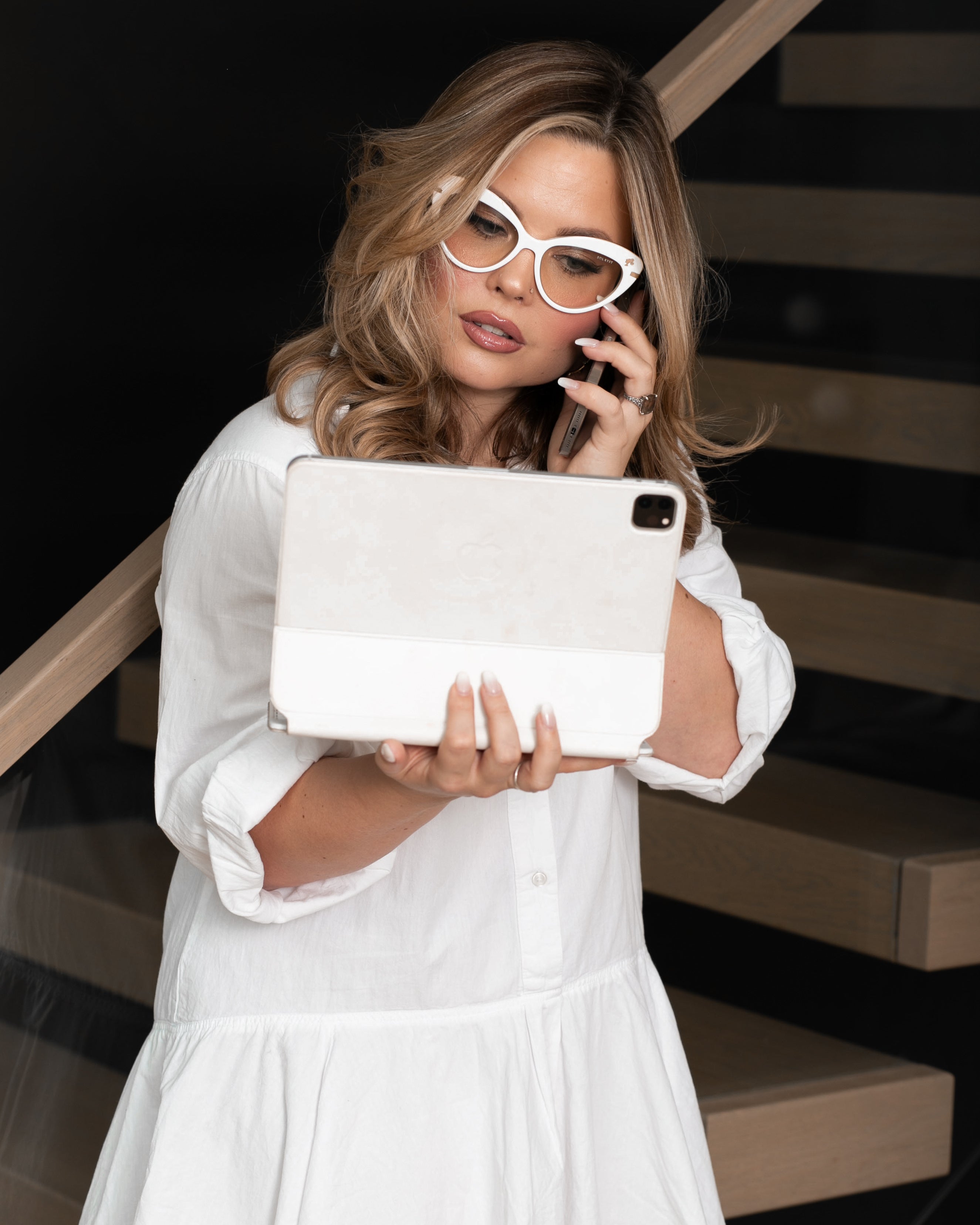 Woman in a white dress holding a tablet and talking on a phone, standing on a staircase.