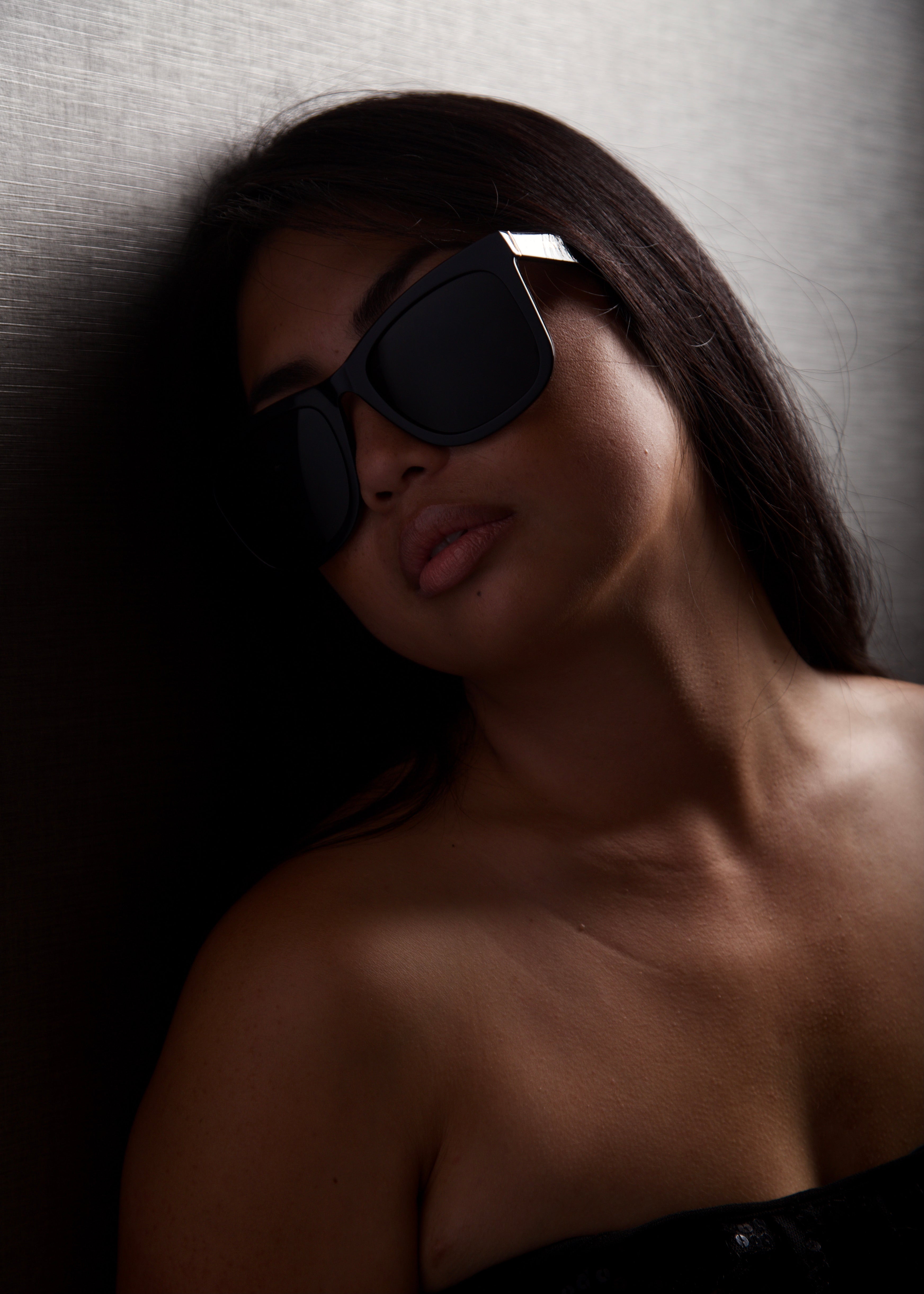 Woman wearing oversized black sunglasses with dark lenses, leaning against a textured wall in dim lighting.