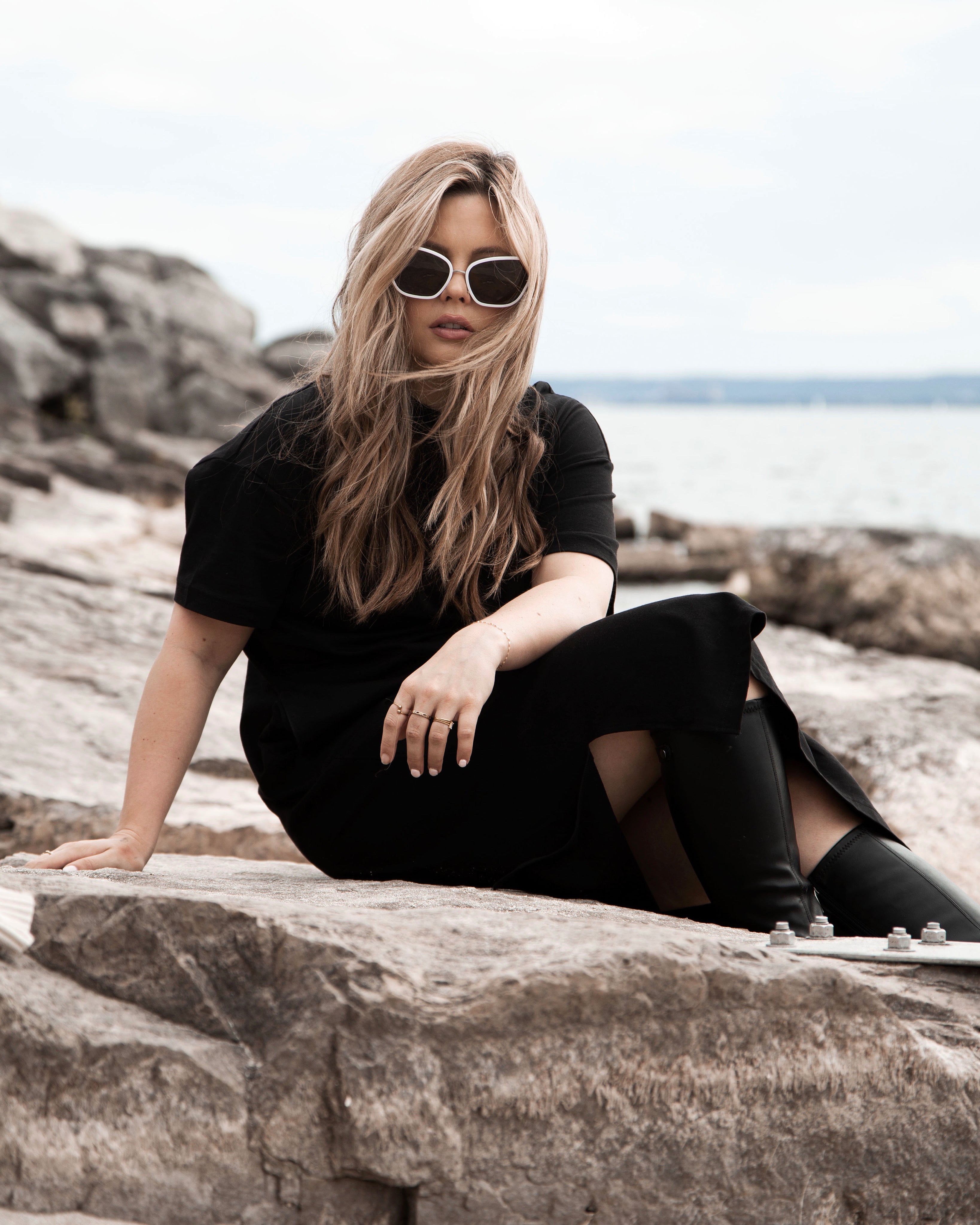 Model sitting on rocks near a lake, wearing a black dress and womens cat eye sunglasses.